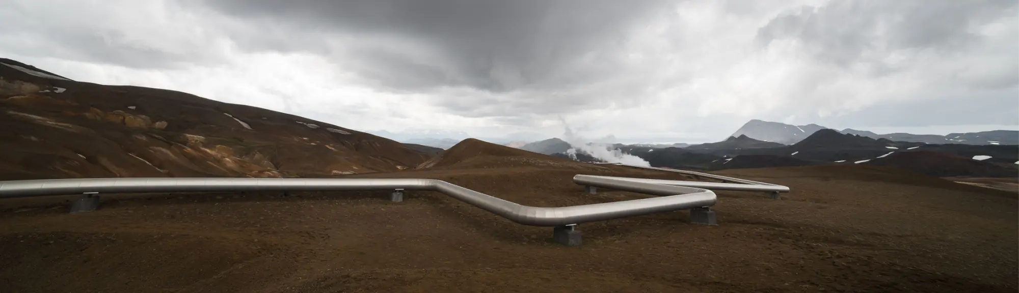 Geothermal pipeline system running across barren terrain with steam rising in the background.