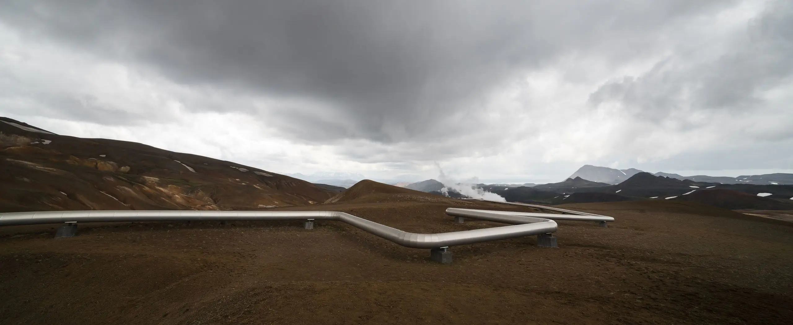 Geothermal pipeline system running across barren terrain with steam rising in the background.