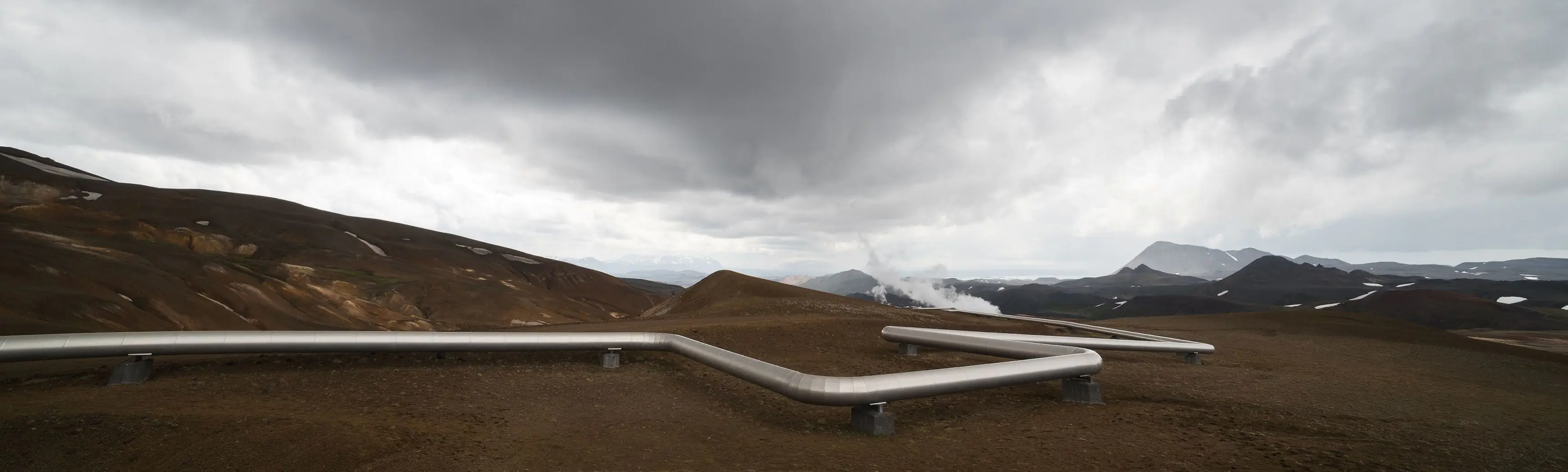 Geothermal pipeline system running across barren terrain with steam rising in the background.